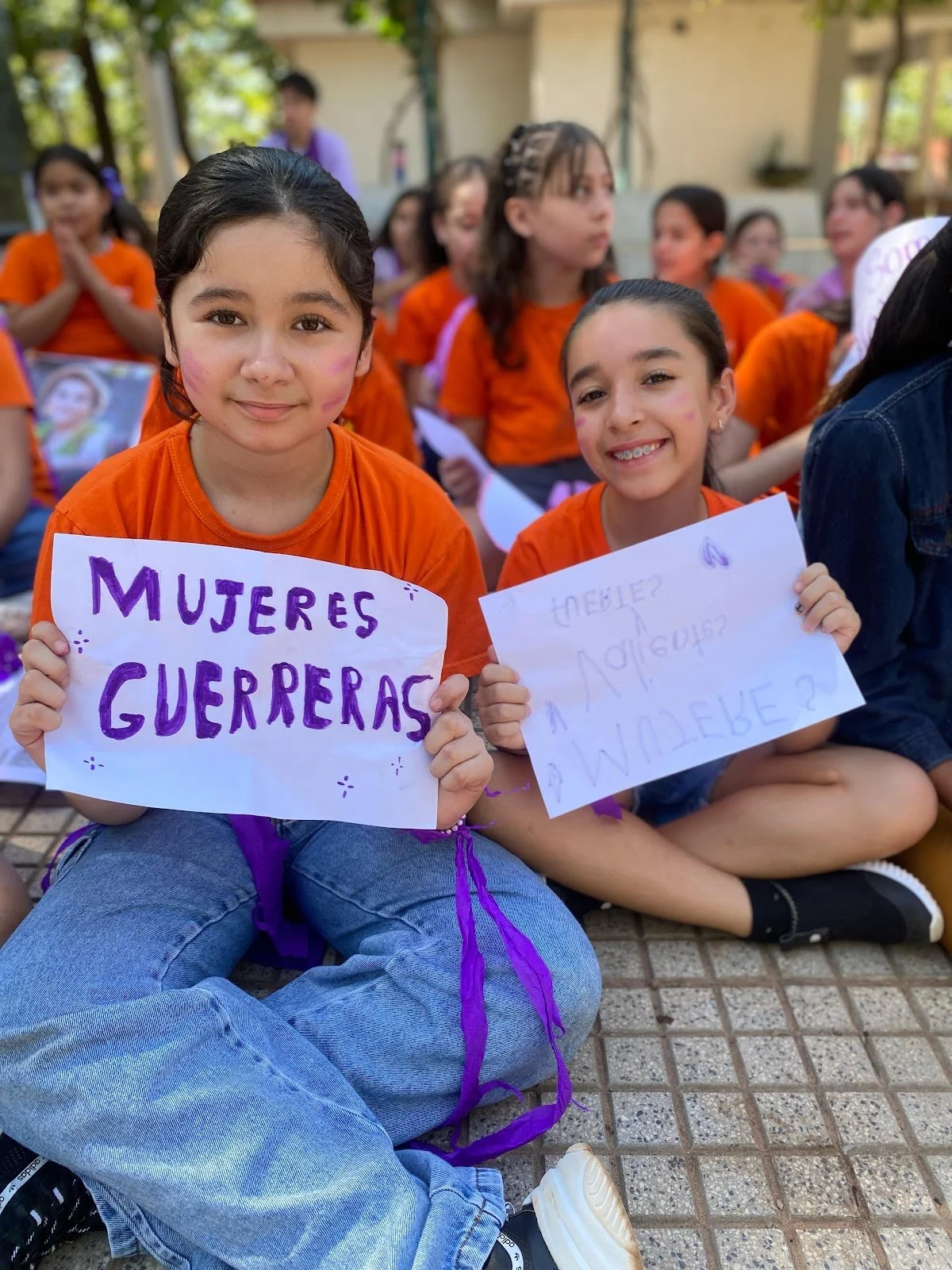 Young girls sitting outdoors during a protest or rally, holding signs in Spanish, with many wearing orange shirts and smiling.