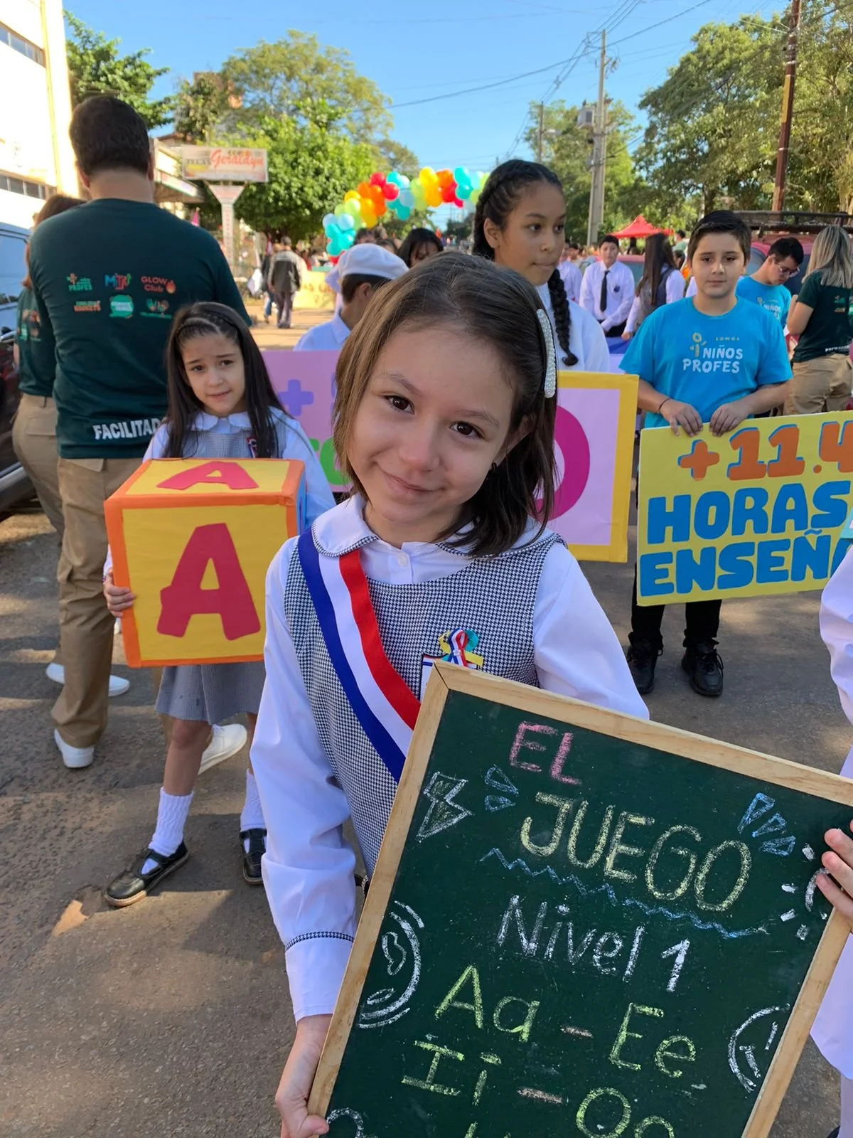 Young girl holding a blackboard with writing, standing among other children participating in a parade or celebration event outdoors. The children are holding colorful signs and banners, and there is a balloon arch in the background.