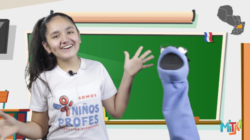 A young girl smiling and gesturing with her hand, standing next to a puppet in front of a classroom green chalkboard.