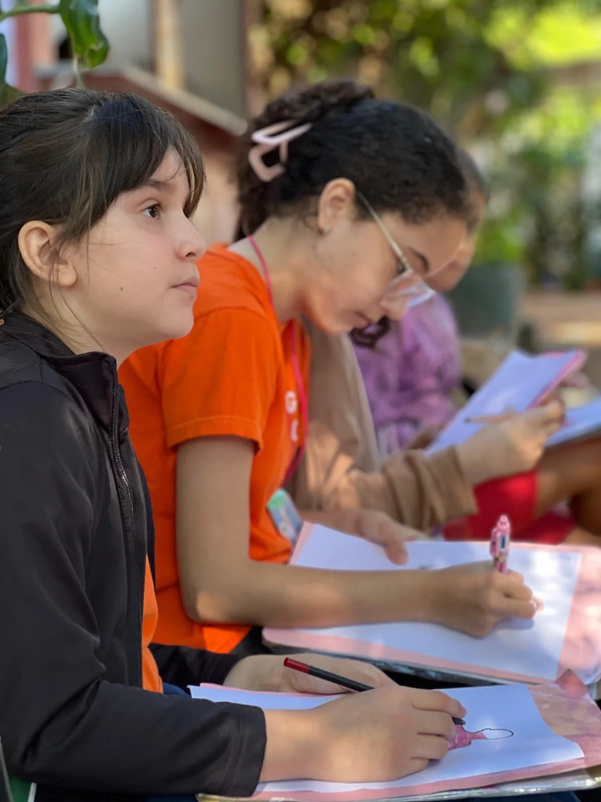 Children sitting in a row outdoors, engaged in drawing or writing on paper.