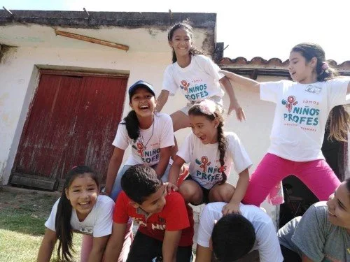Group of children playing and laughing outdoors on grass, with a building in the background.