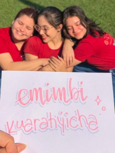 Three young women in red shirts smiling and hugging each other outdoors, holding a sign that reads 'emimbi kuardiyicha' in pink handwriting.