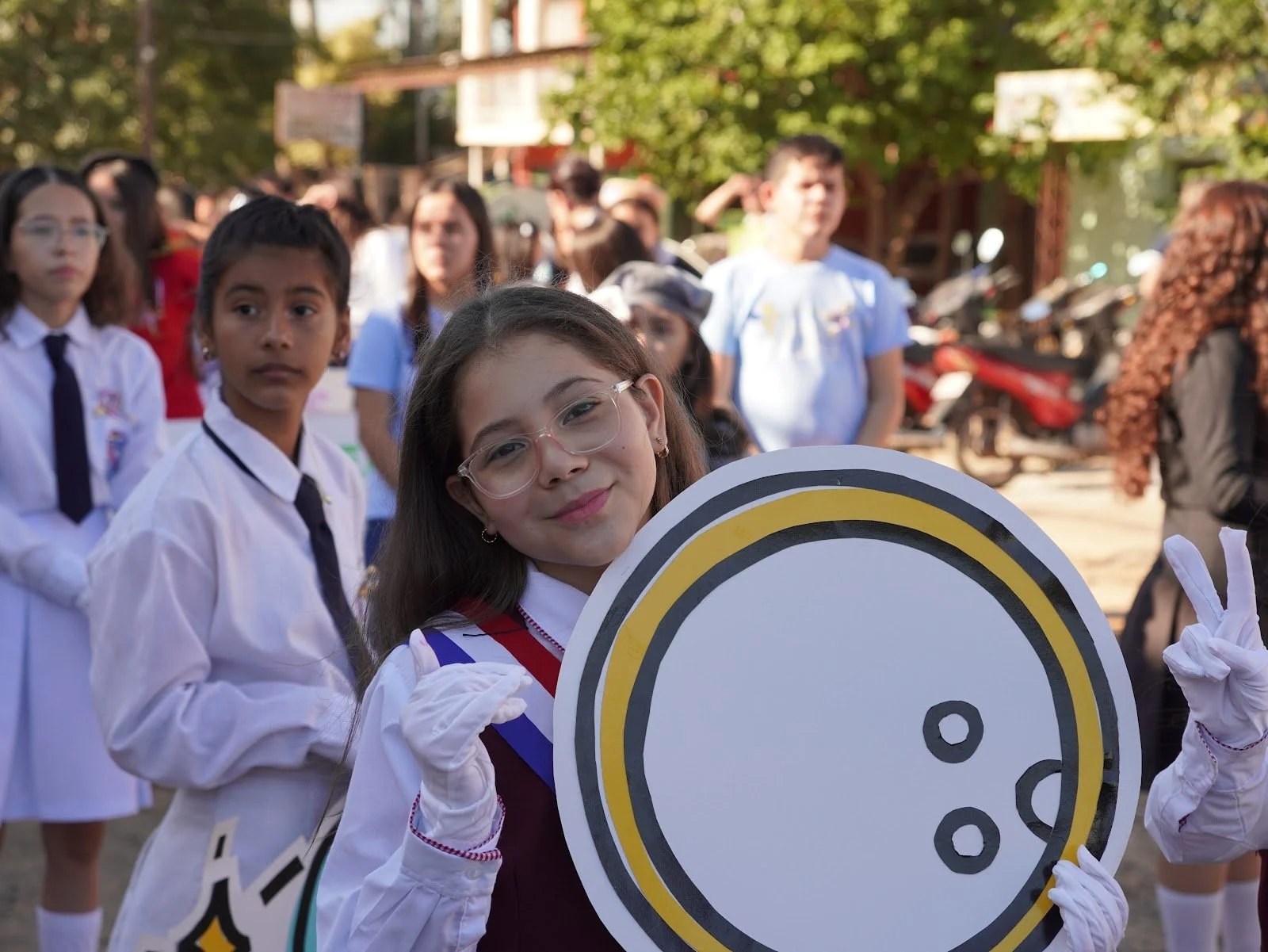 Group of children dressed in white uniforms at an outdoor event, with one girl in the foreground smiling and holding a large cutout of a food plate, with a crowd and trees in the background.