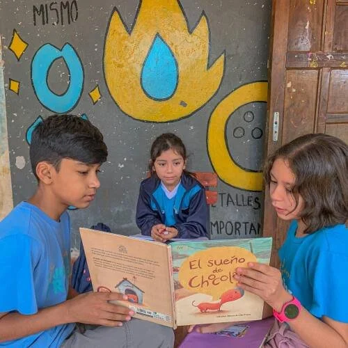 Three children in blue uniforms sitting in a classroom, with one girl reading a book titled "El Sueño de Chocolate" to two other children. The background features colorful cartoon flames, droplets, and shapes on a gray wall.