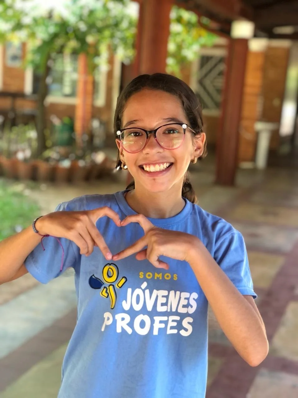 Smiling young girl with glasses making a heart with her hands, wearing a blue T-shirt with the words 'Somos Jóvenes Profes' and a small cartoon character, standing in an outdoor setting with a wooden structure and greenery in the background.