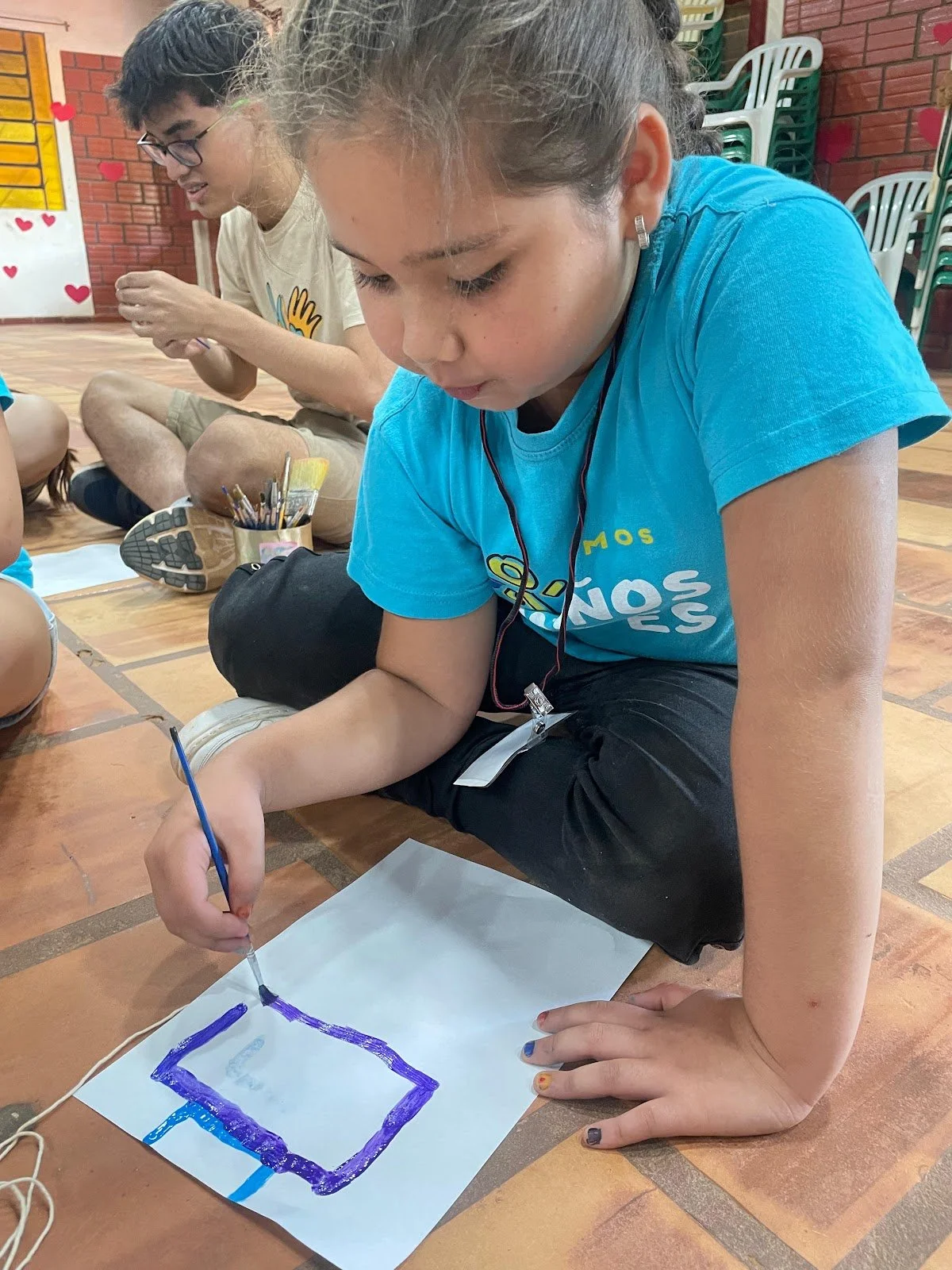 A young girl sitting on the floor, wearing a blue t-shirt and painting a picture with purple and blue colors on a white sheet of paper. Other children are visible in the background, engaging in similar activities in a colorful indoor setting.
