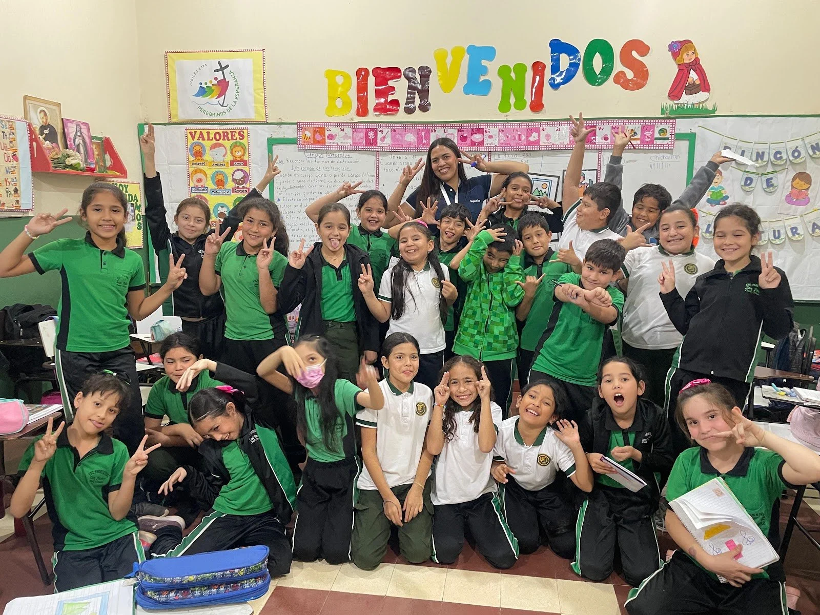Group of school children and a teacher posing in a classroom with colorful decorations and educational posters.
