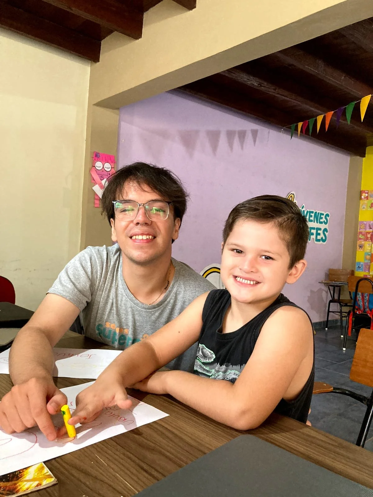 A smiling man with glasses and a young boy with a big smile, sitting at a table with papers and crayons, in a colorful room with decorations in the background.