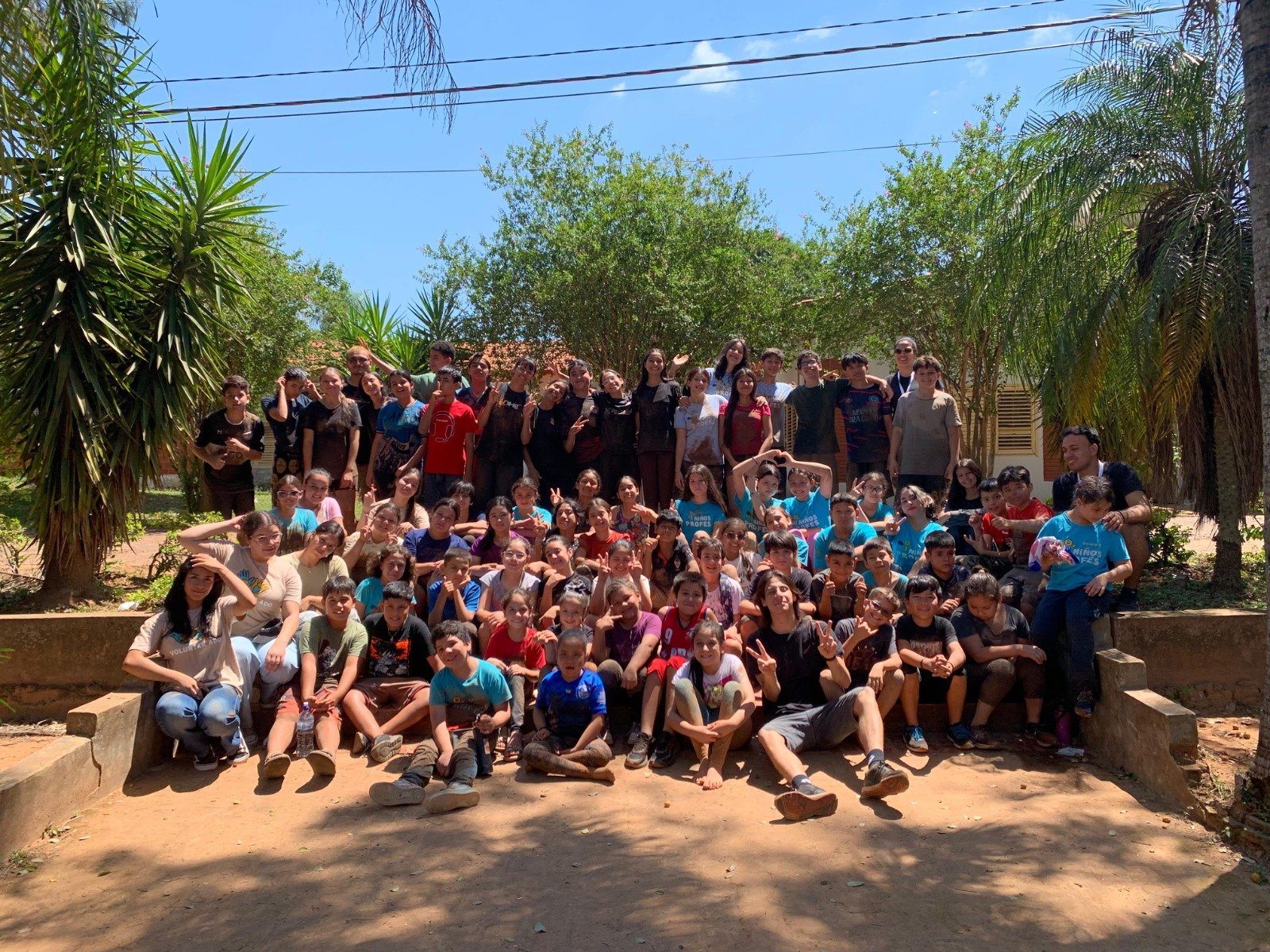 Group of children and teenagers outdoors on a sunny day, posing for a group photo with trees and blue sky in the background.