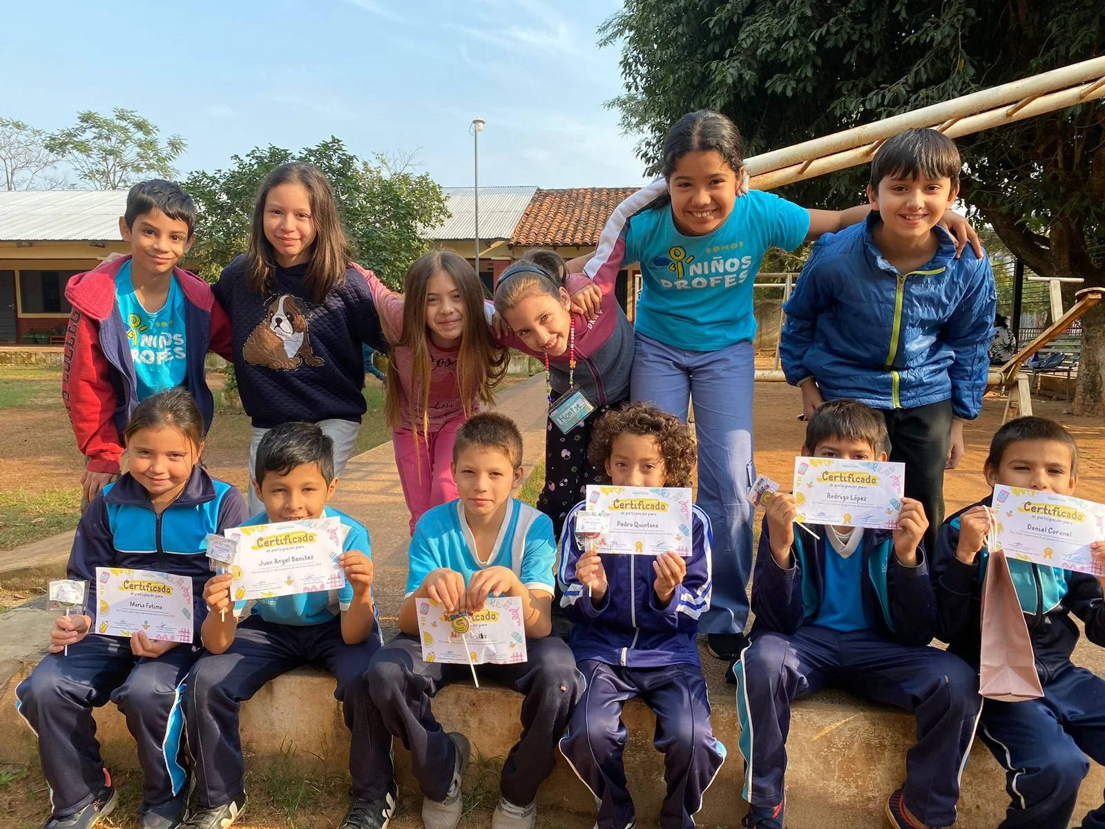 Group of children outdoors, posing with certificates, smiling, some kneeling, others standing, in a schoolyard with trees and buildings in the background.
