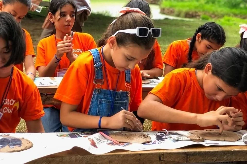 Group of young girls in orange t-shirts working on clay art projects outdoors at a long table.