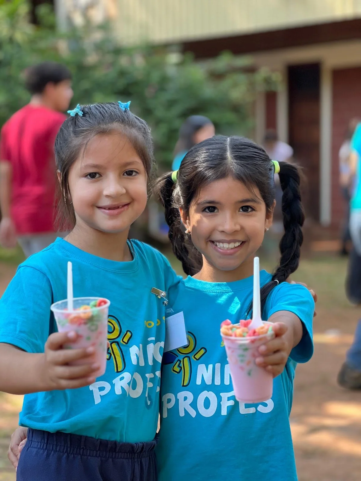 Two young girls smiling and holding cups of pink frozen treat with colorful cereal toppings at outdoor event.