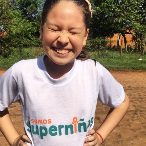 Young girl smiling outdoors on a sunny day, wearing a white t-shirt with colorful text, standing on a dirt field with trees and a fence in the background.