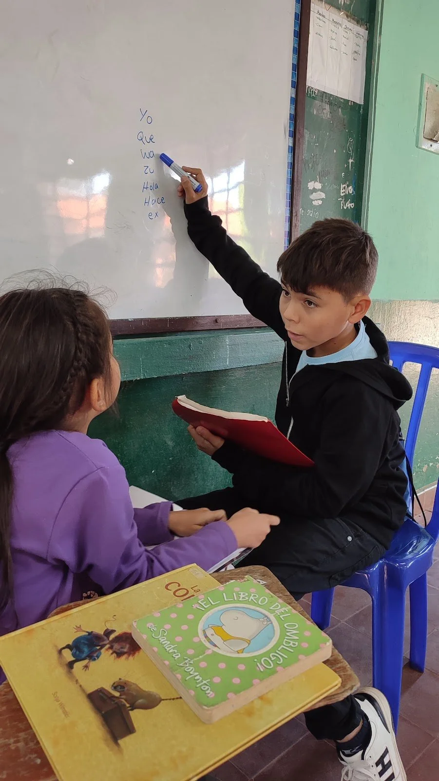 A classroom scene with a boy writing on a whiteboard and a girl sitting and reading a book. The boy is holding a red folder, and there are books on the desk, including one titled 'Il Libro Dell'Ombelico' featuring a cartoon whale.