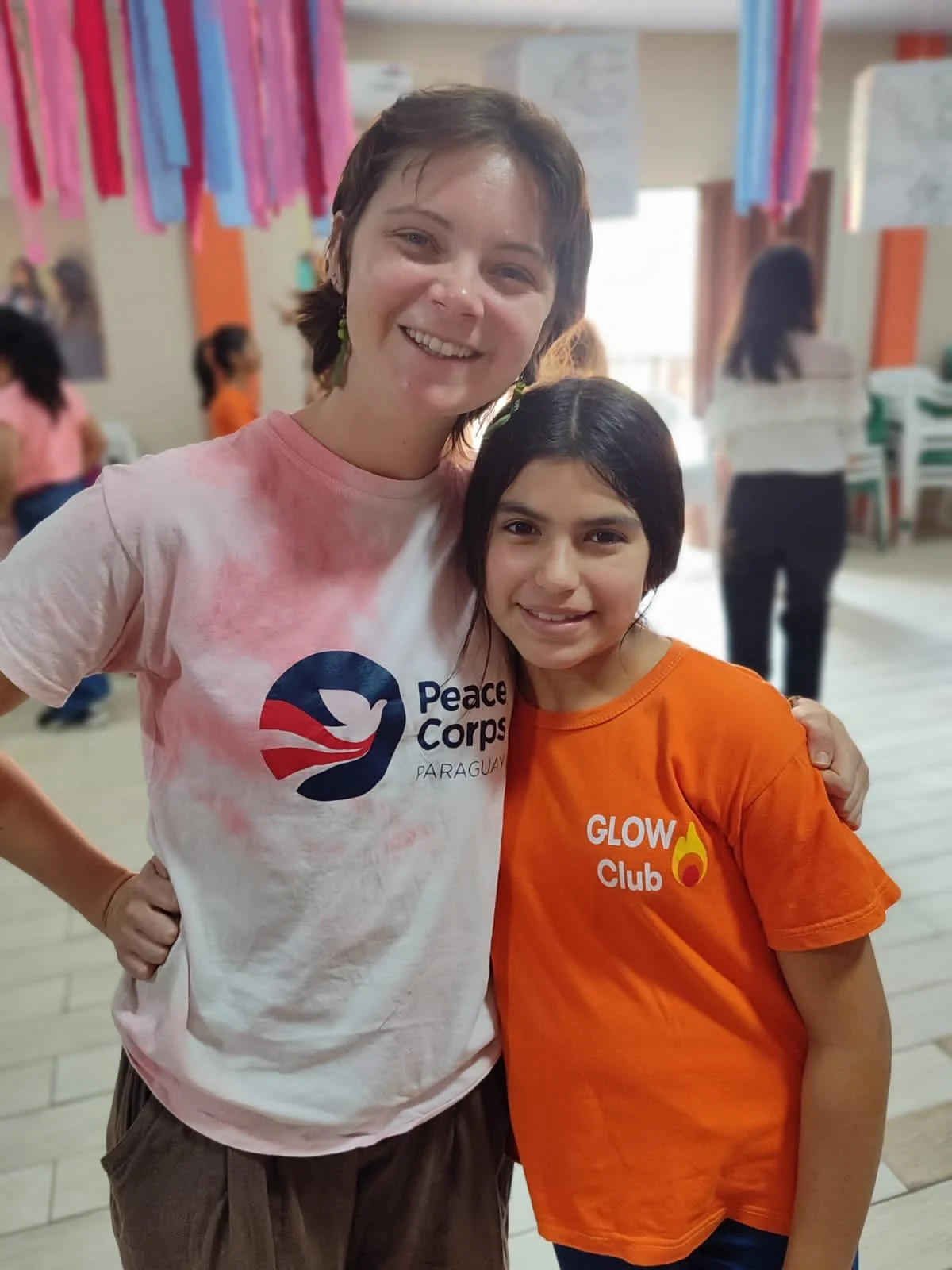 Two smiling females, one adult and one young girl, standing arm in arm indoors at an event. The adult wears a white T-shirt with a Peace Corps Paraguay logo, and the girl wears an orange T-shirt with 'GLOW Club' and a flame graphic. Background featur