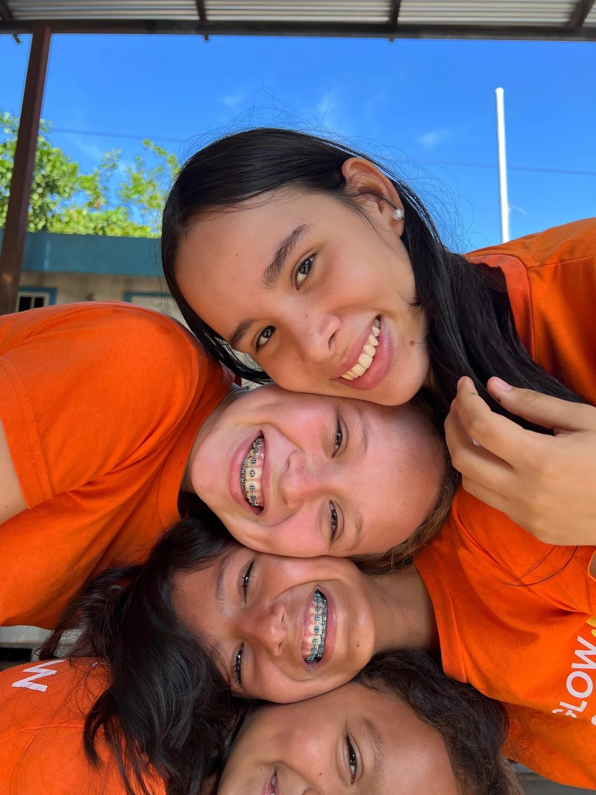 Group of five smiling young women in orange shirts, lying close together outdoors with blue sky and green trees in the background.
