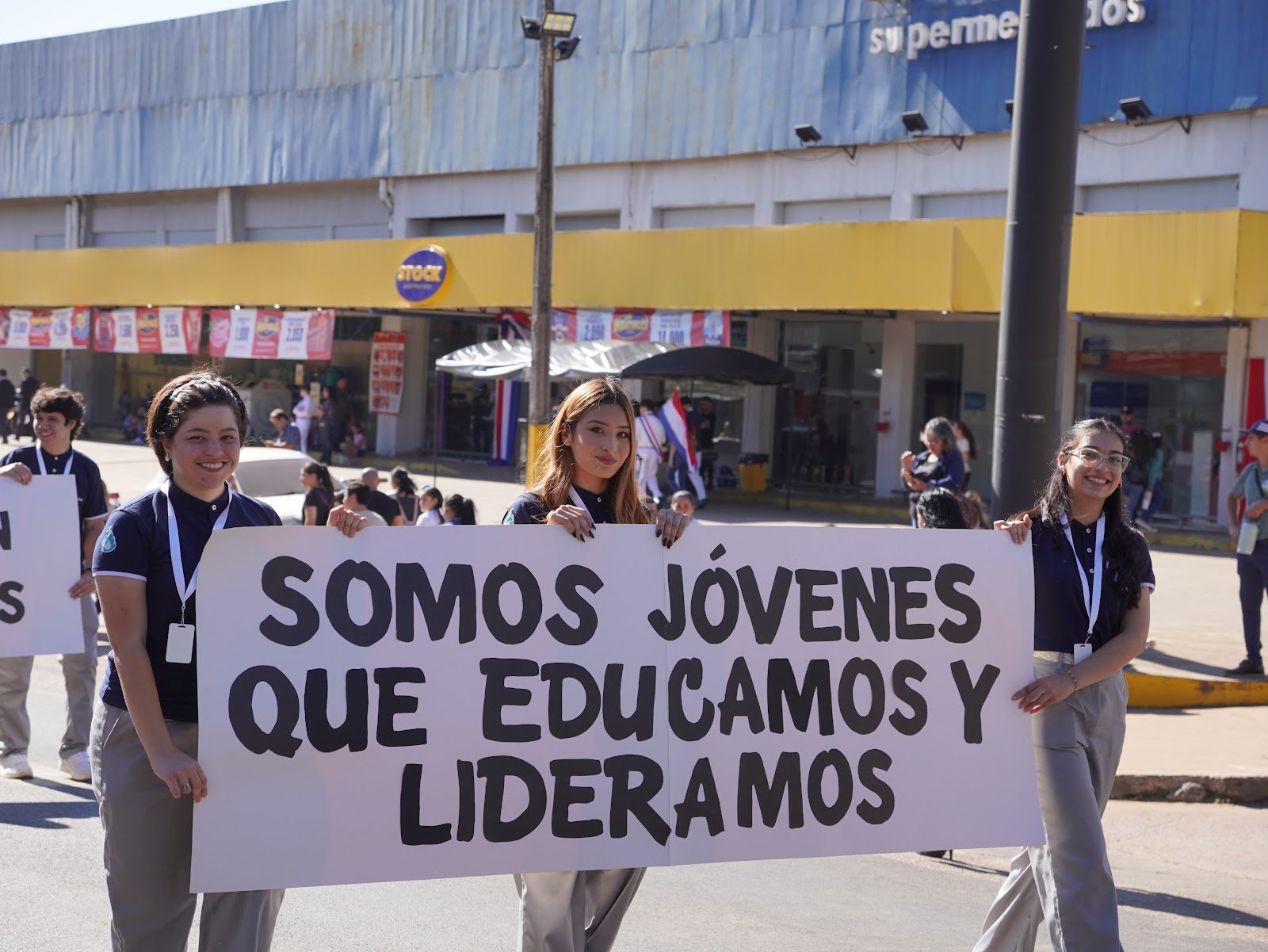 Young women participating in a march or protest, holding a sign that reads 'SOMOS JÓVENES QUE EDUCAMOS Y LIDERAMOS'. They are outdoors in front of a store with a yellow sign and a crowd of people in the background.