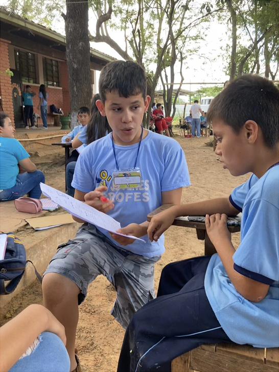 Two boys in blue T-shirts sitting outdoors, engaged in a conversation and reviewing papers, with other children and adults in the background.