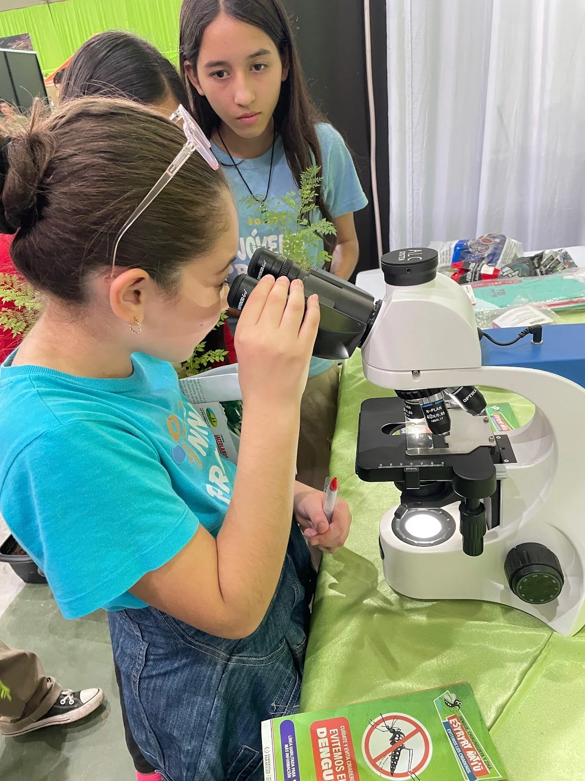Five children observing a microscope at an educational exhibit, with insect-related signage and materials on the table.