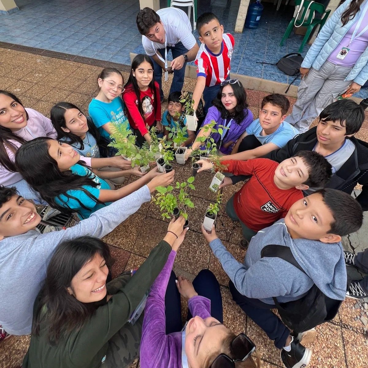 Group of children and a few adults gathered around small potted plants on a table, participating in a planting activity.