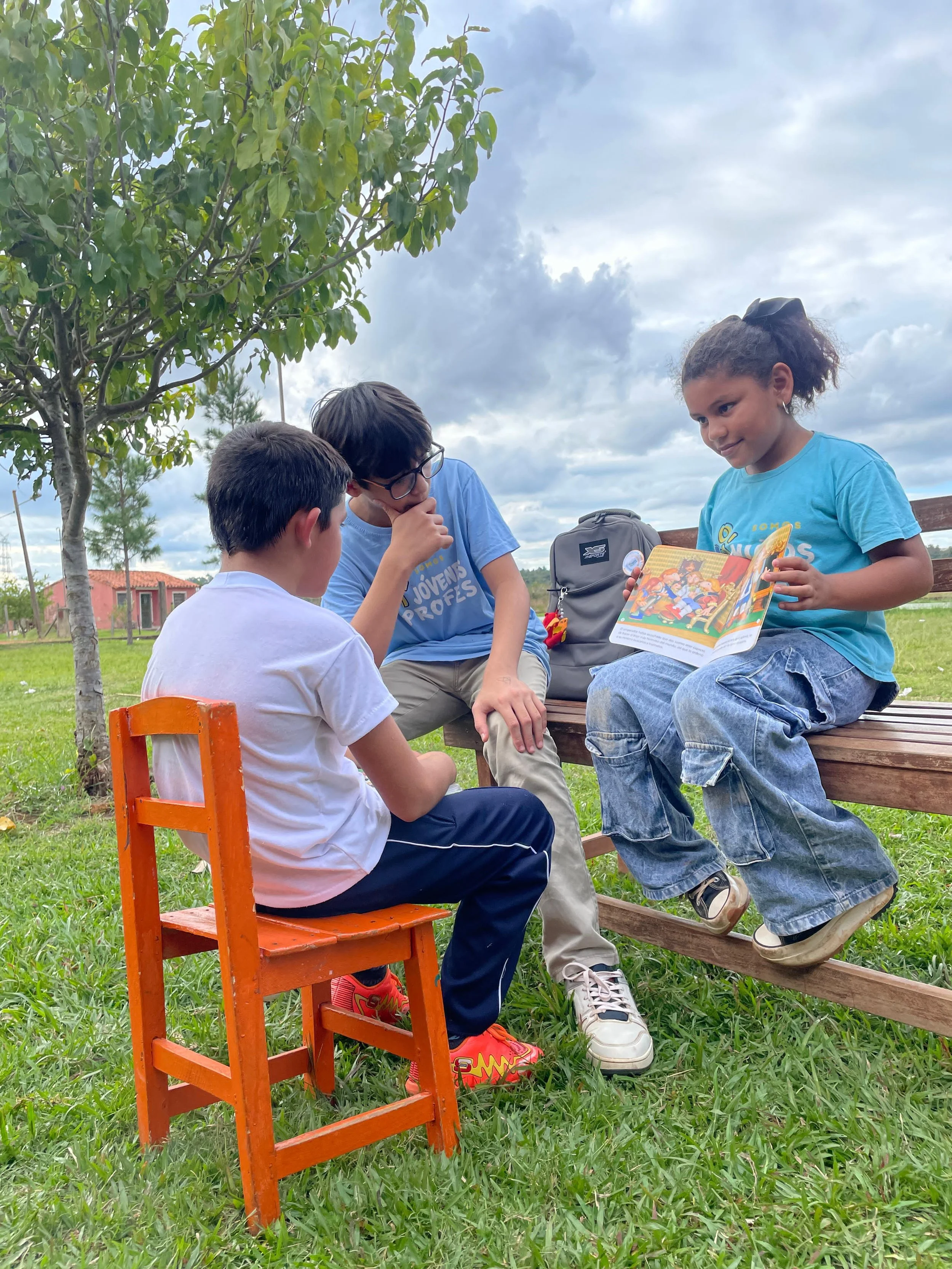 Three children sitting outside on grass, reading a colorful storybook together under cloudy sky, with trees and houses in the background.