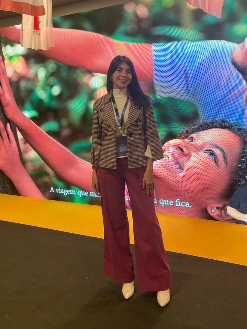 A woman with dark hair wearing a beige turtleneck, checkered blazer, maroon wide-leg pants, and white boots standing in front of a colorful large digital display with abstract images and partial Portuguese text.