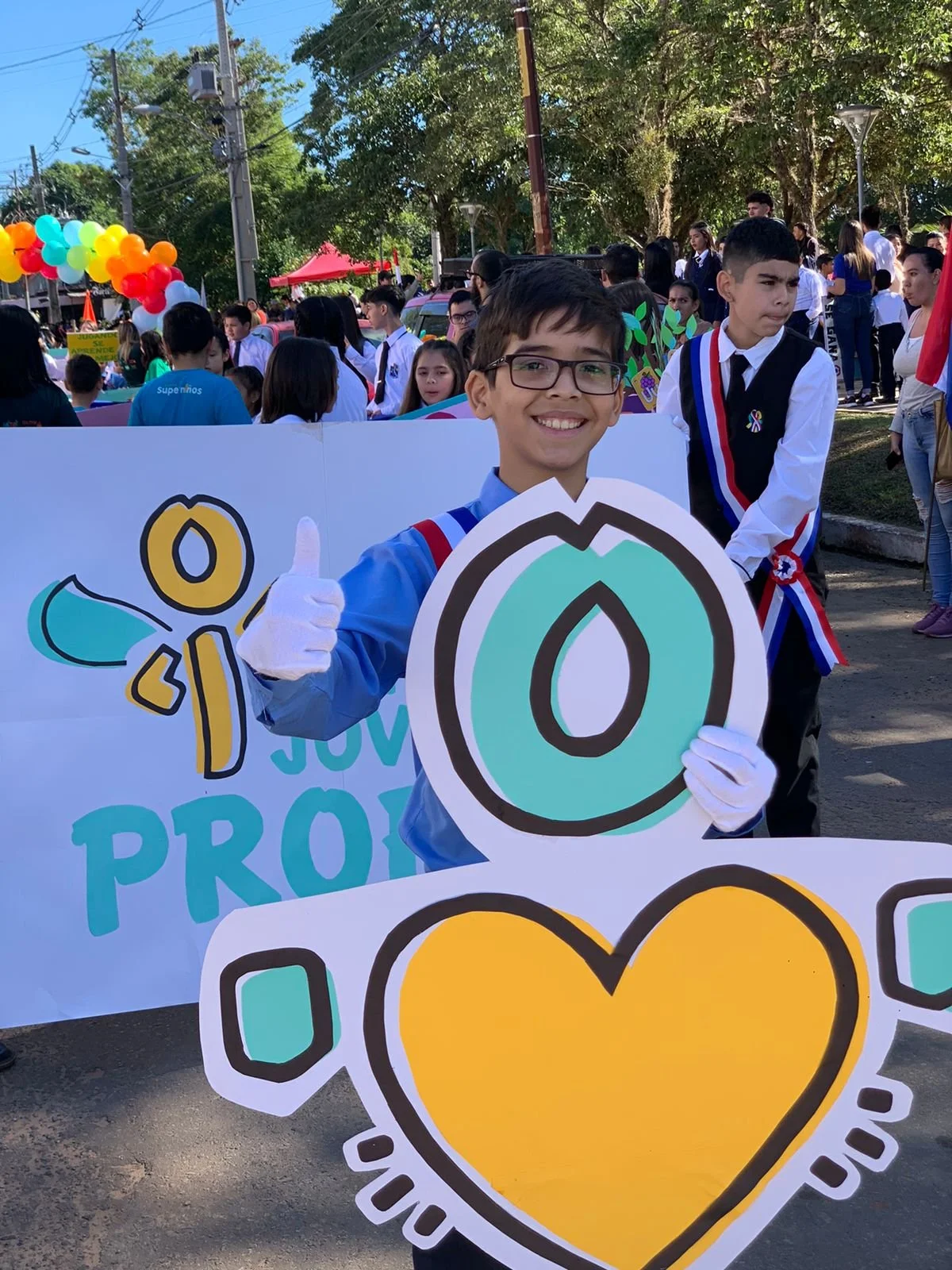 Smiling young boy wearing glasses and white gloves holding a large sign with a colorful heart design during a parade or rally, surrounded by other young participants and onlookers.