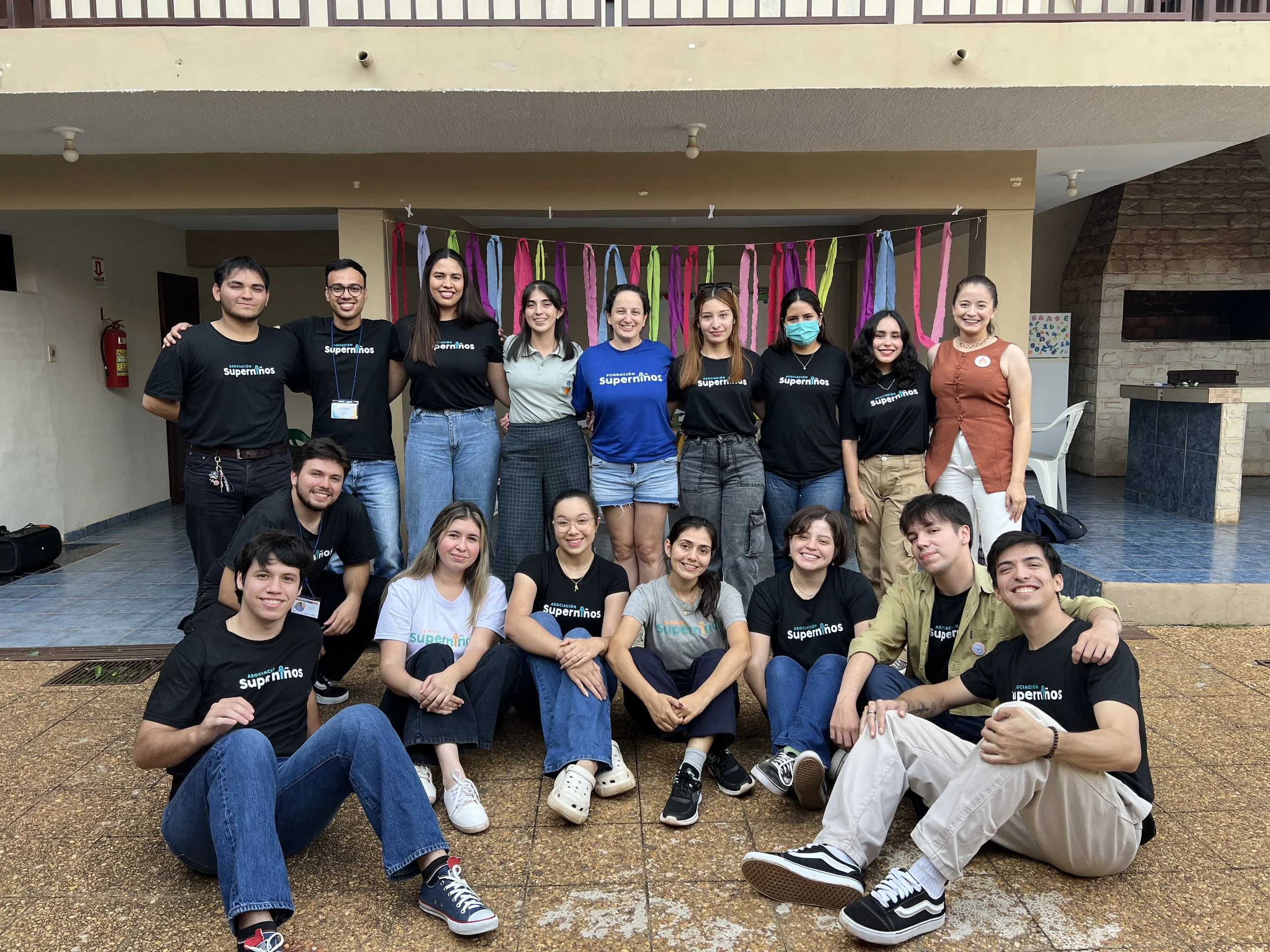 A group of young people posing for a photo indoors, some wearing black T-shirts with the logo of 'Asociación Superniños', smiling and standing in front of colorful fabric streamers hanging from a line.