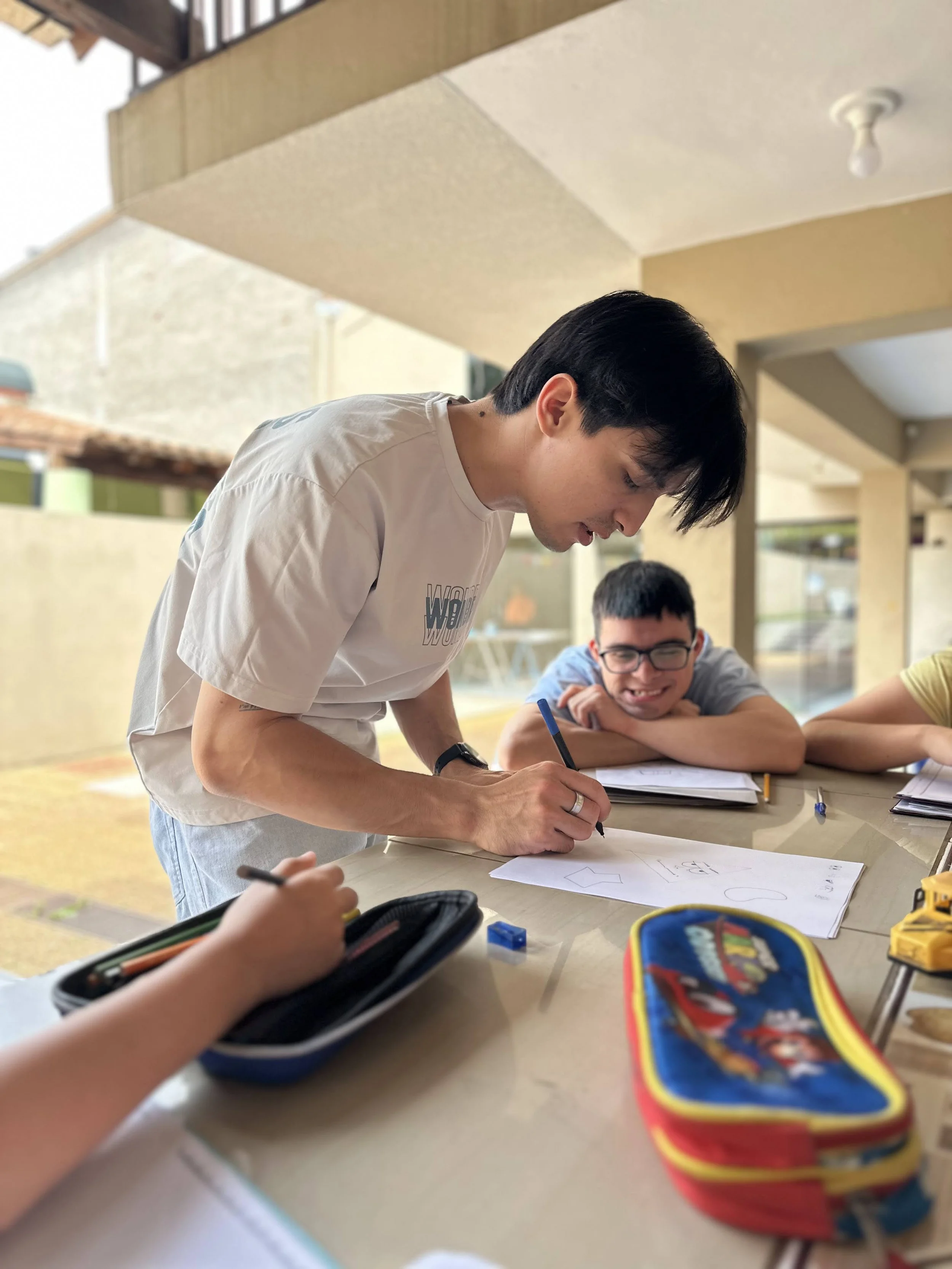 A young man with dark hair wearing a white T-shirt leans over a table, writing or drawing on a piece of paper, while smiling and looking at someone across the table. In the background, another person with glasses and a blue shirt is smiling. The setting appears to be a casual indoor or semi-outdoor space with a beige wall and large windows.