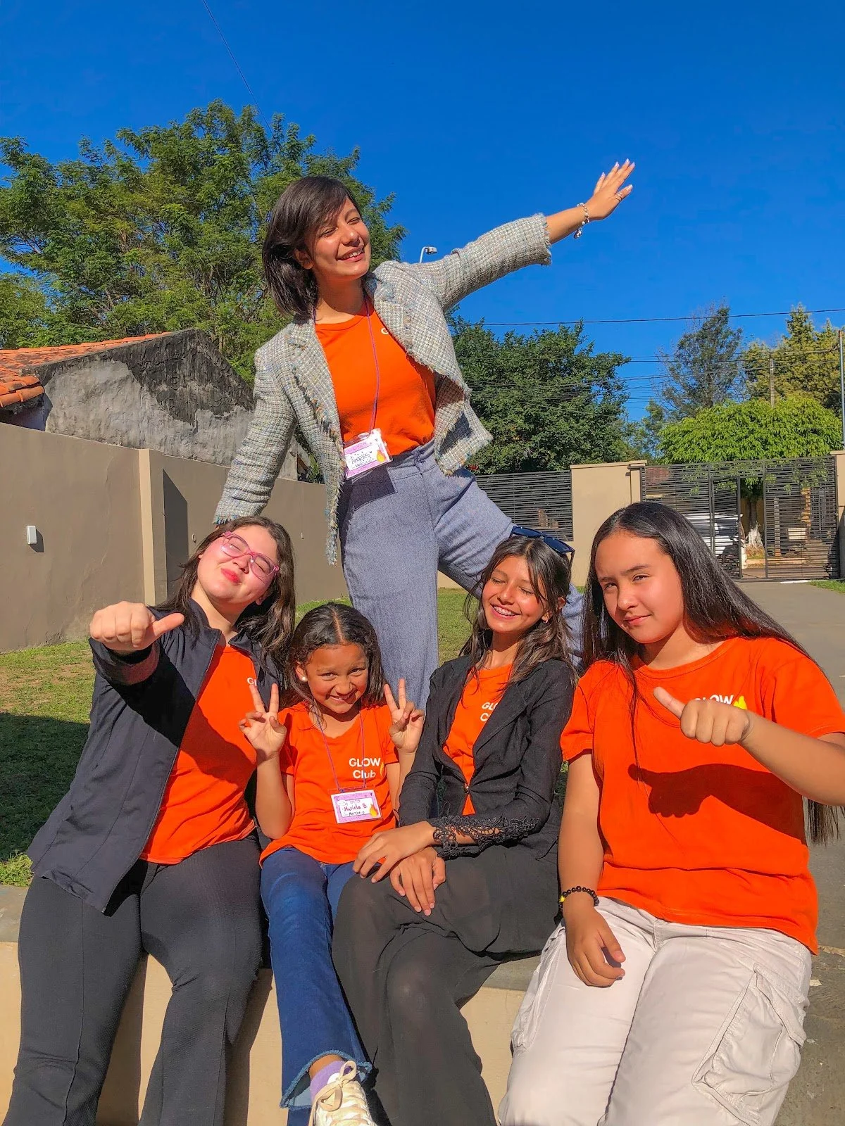 A group of five women, four young girls and one adult woman, enjoying outdoors on a sunny day, smiling and posing for the camera, with a bright blue sky and green trees in the background.