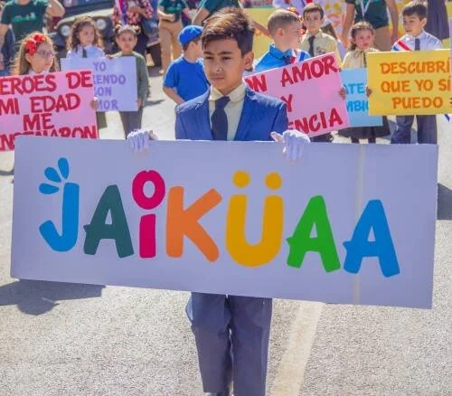 A young boy in a suit holding a large sign that spells 'JAIKUALA' in colorful letters, surrounded by children and adults holding signs in Spanish during a parade or demonstration.