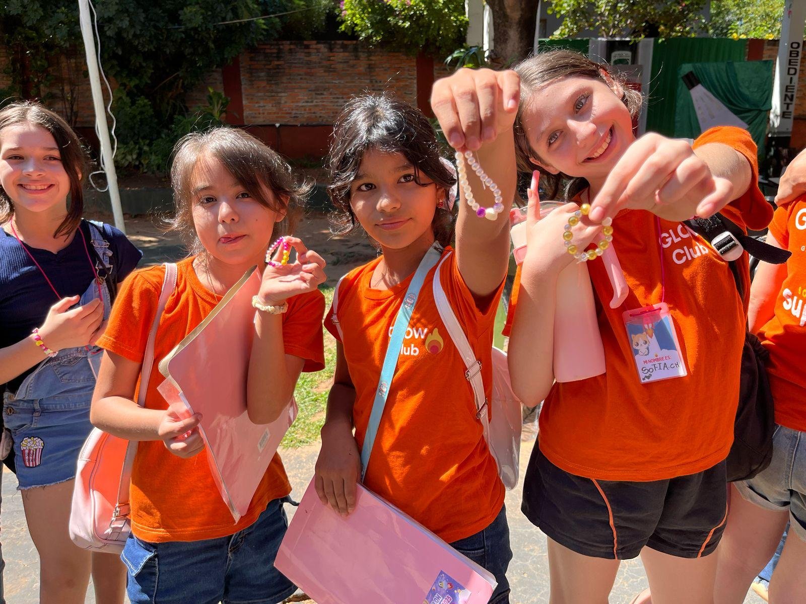 Group of kids holding beaded jewelry and pink folders outside in the sun, smiling.