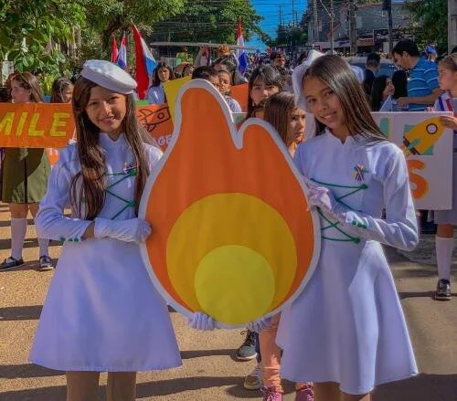 Two young girls in white dresses holding a large flame-shaped sign during a parade or celebration.