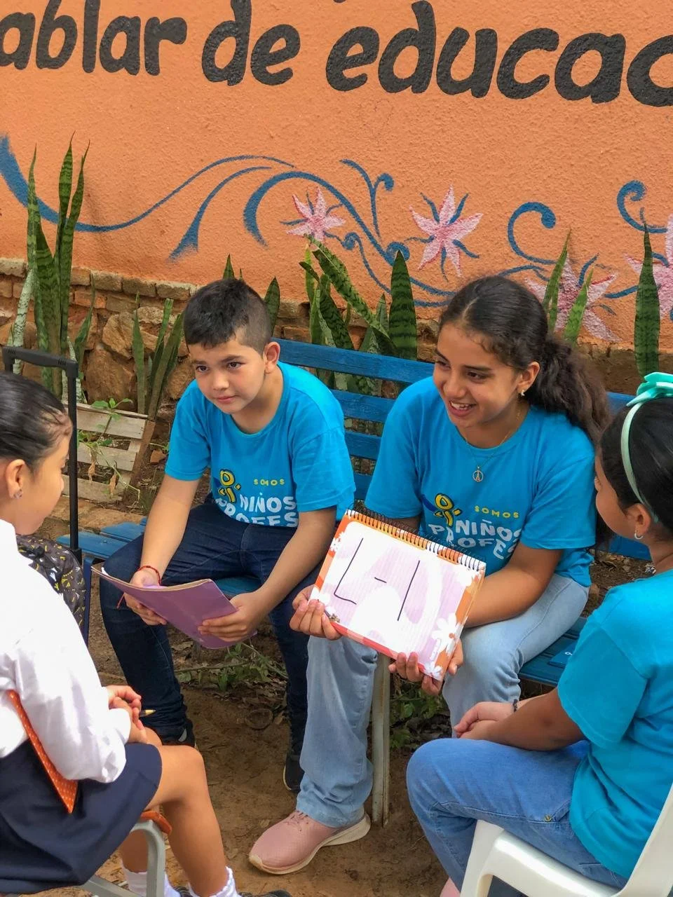 Group of children and young adults participating in an outdoor educational activity, with two children holding notebooks and engaging with the others, sitting on benches in front of a vibrant orange wall with floral and decorative artwork.