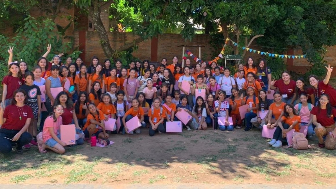 Group of children and adults outdoors, some holding pink folders or bags, in front of trees and a brick wall, during daytime.