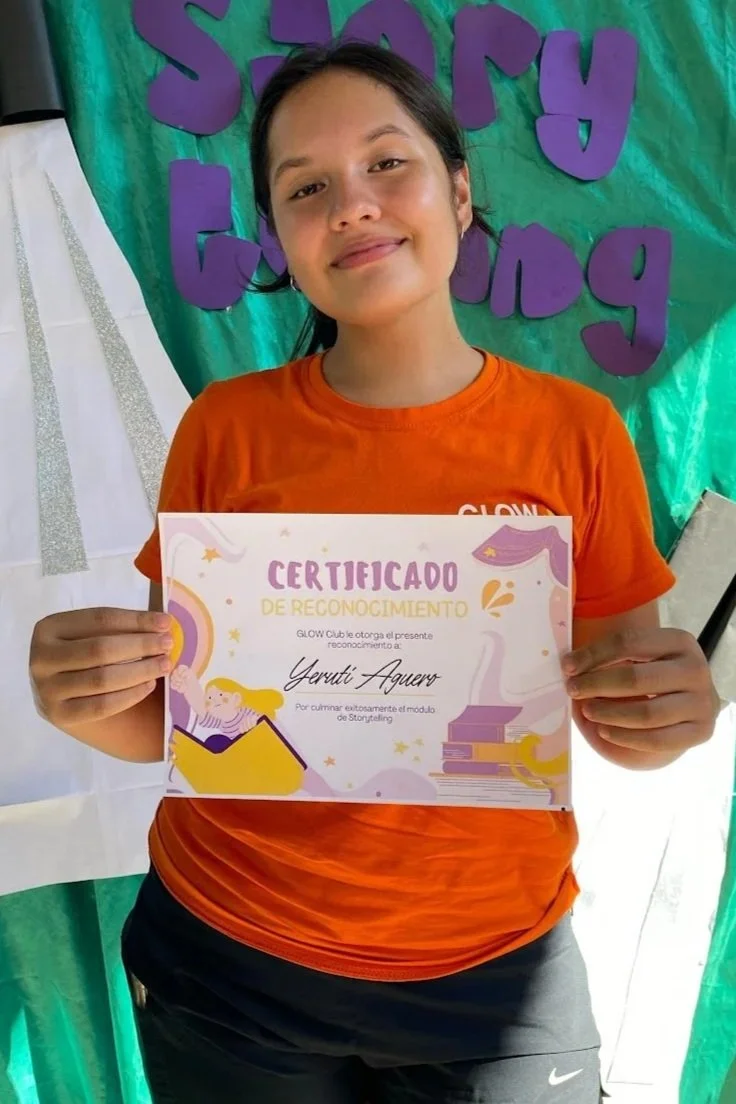 A young girl holding a certificate of recognition, standing in front of a colorful backdrop with purple letters spelling 'Story'. She is smiling and wearing an orange shirt.