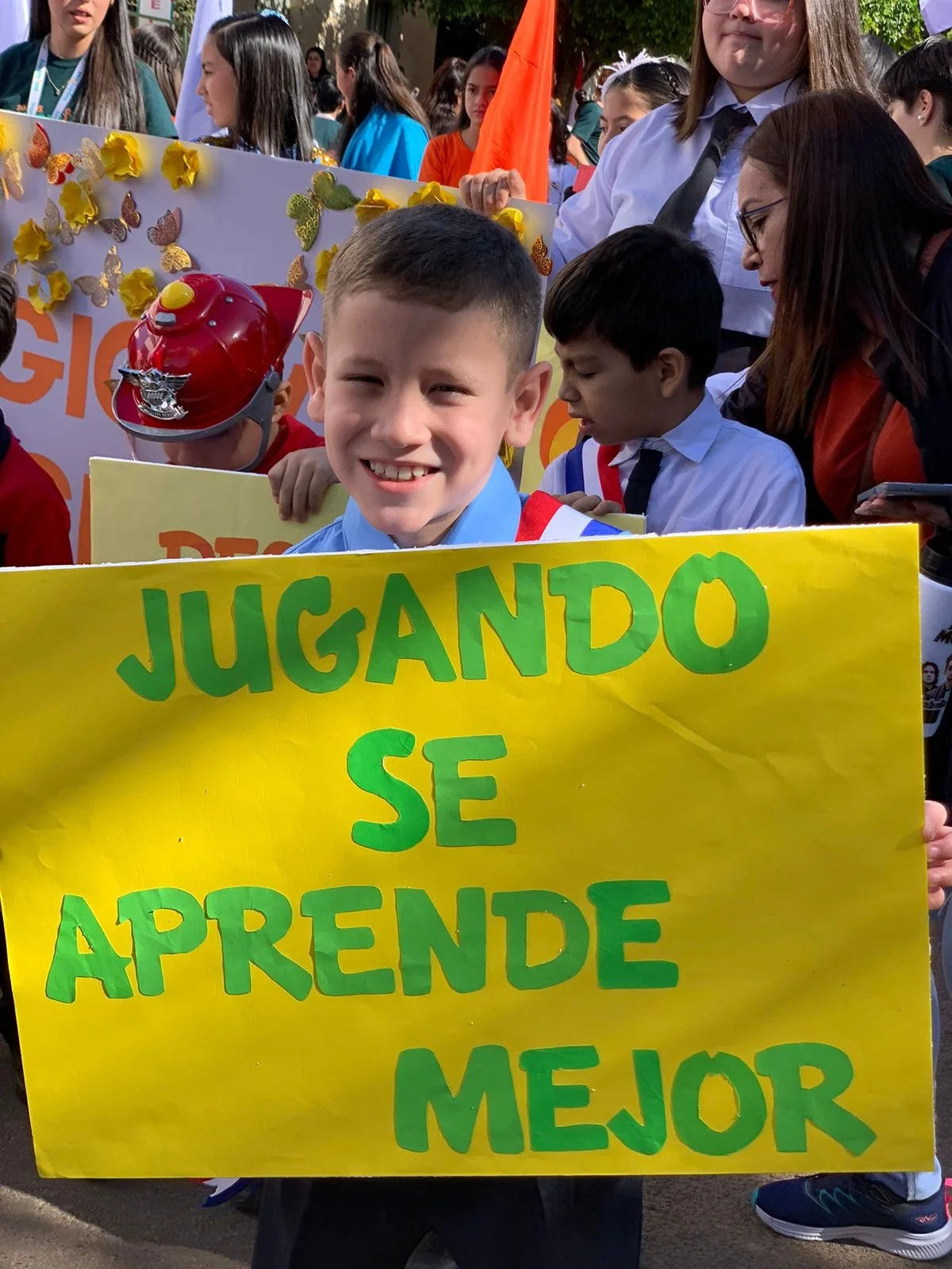 Young boy holding a yellow sign with green letters that reads, "Jugando se aprende mejor," during a parade or public event with other children and adults in the background.