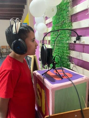 A young boy wearing headphones is speaking into a microphone at a booth, with green foliage and balloons in the background.