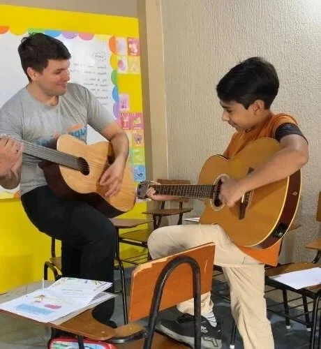 Two boys playing acoustic guitars in a classroom with chairs and a colorful background