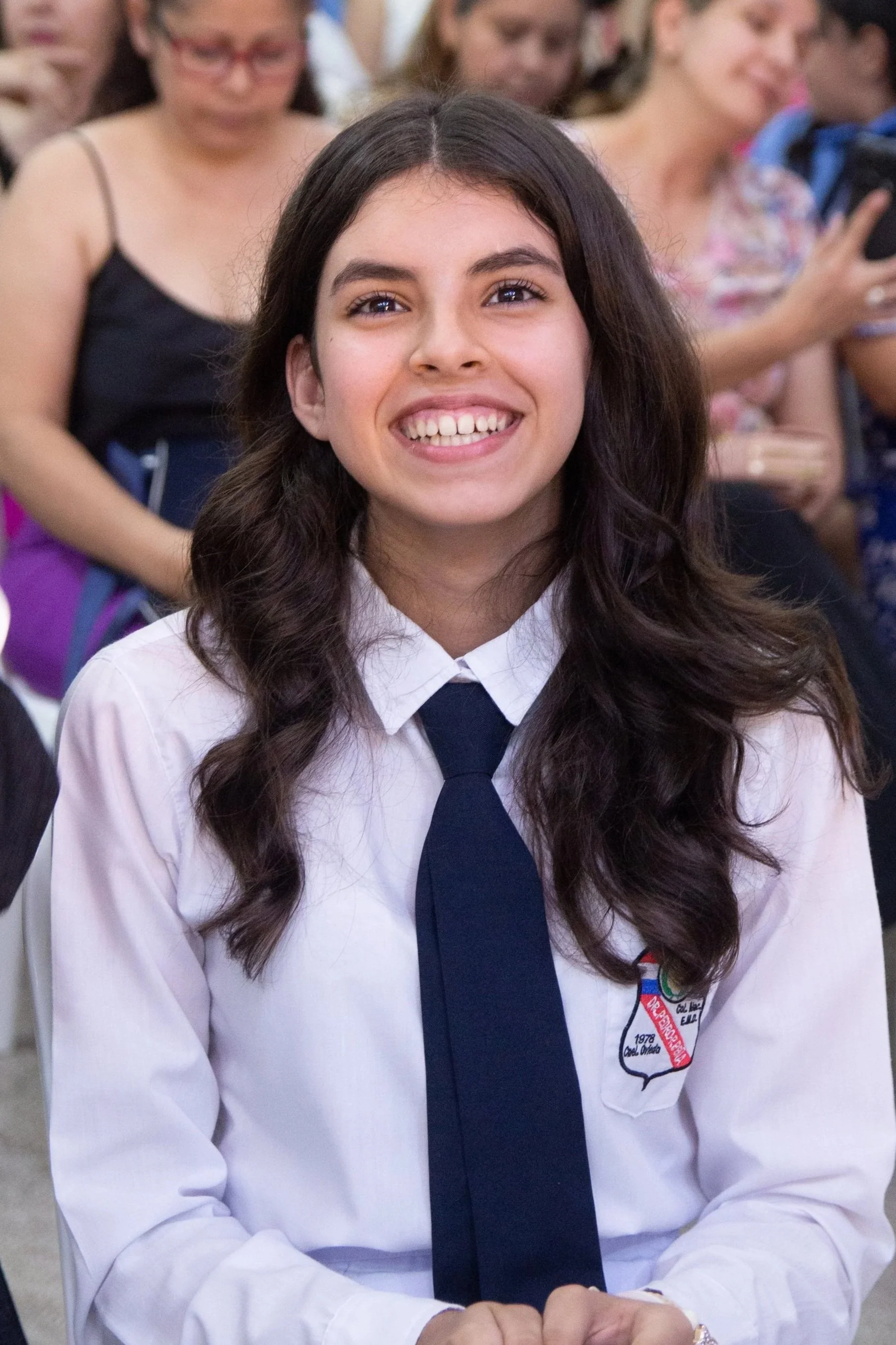 Smiling young woman with long dark hair wearing a white shirt and dark tie, sitting at an event with people in the background.