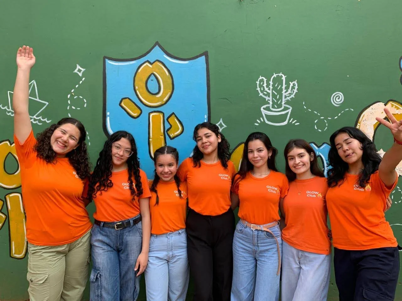 Group of seven women standing in front of a green wall with colorful graffiti and designs, all smiling and wearing matching orange T-shirts that say 'GLOW CLUB'.