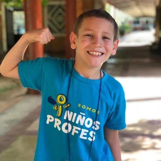 A young boy smiling and flexing his right arm outdoors, wearing a blue T-shirt with a playful design and text.