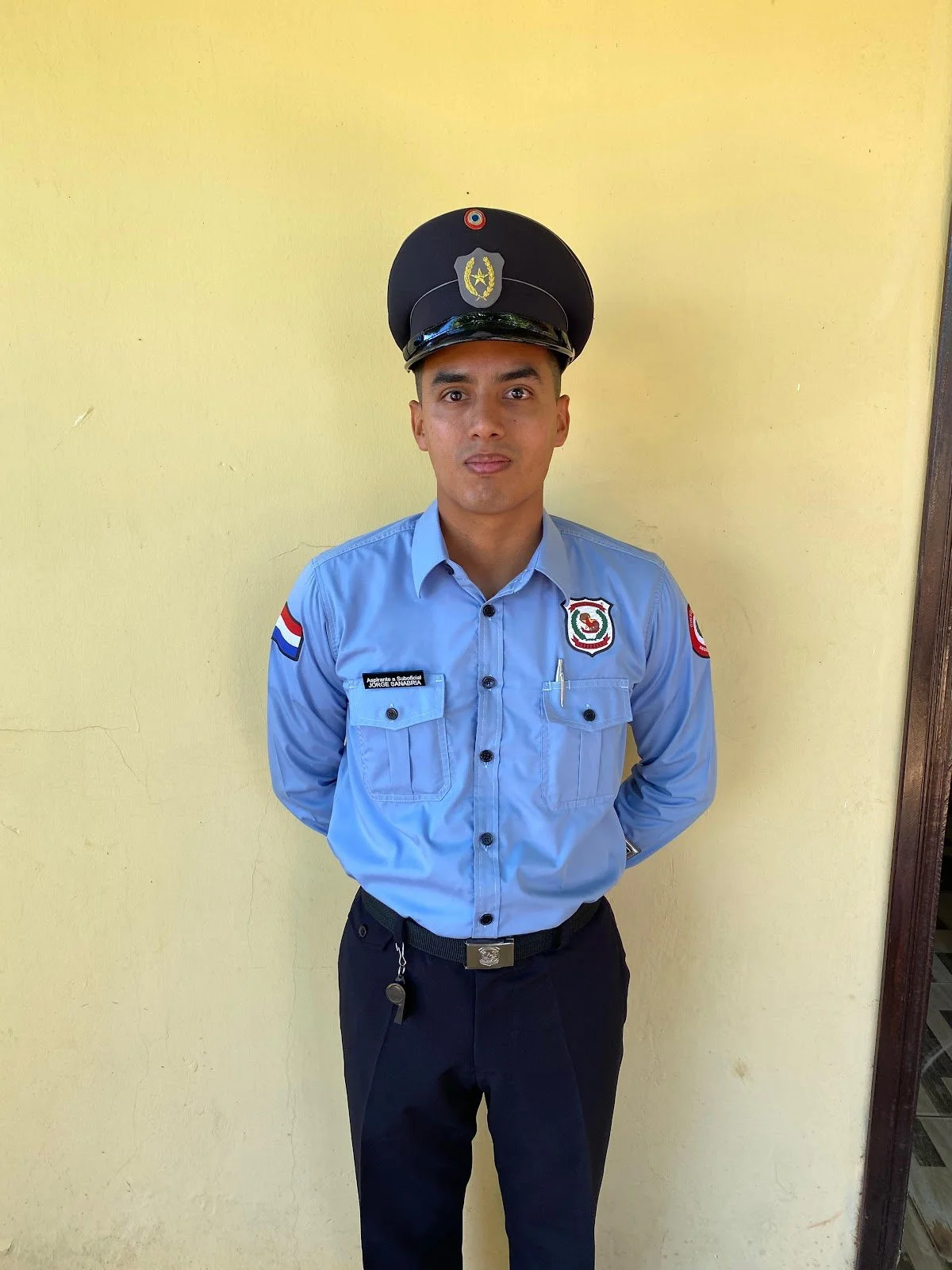 A young male security officer in uniform standing against a yellow wall, wearing a light blue shirt with patches, dark pants, and a black cap with a badge.
