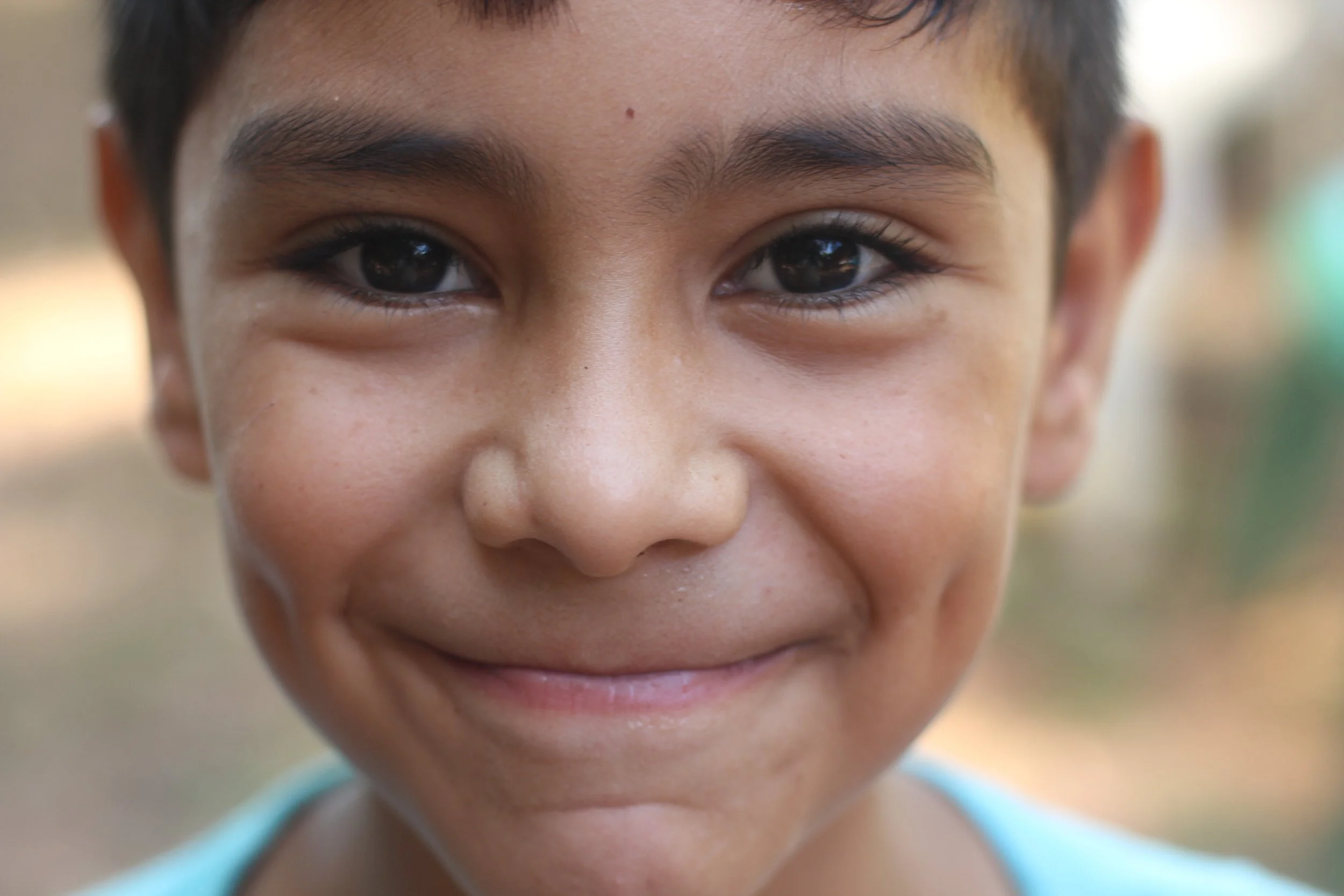 Close-up of a young boy's smiling face showing part of his eyes, nose, and mouth outdoors with soft blurred background.