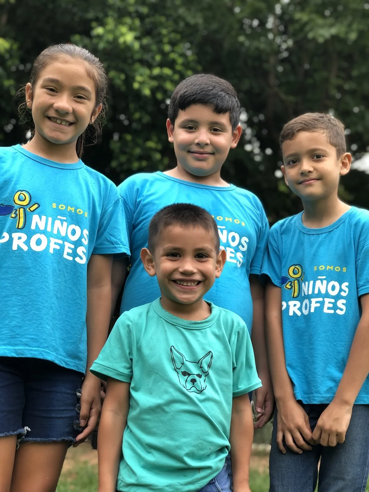 Group of four children smiling outdoors, wearing blue shirts with Spanish text, standing in front of green trees.