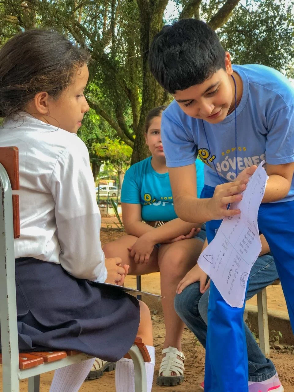 A group of young people outdoors, with one boy showing a paper to a girl while two others watch, surrounded by trees.