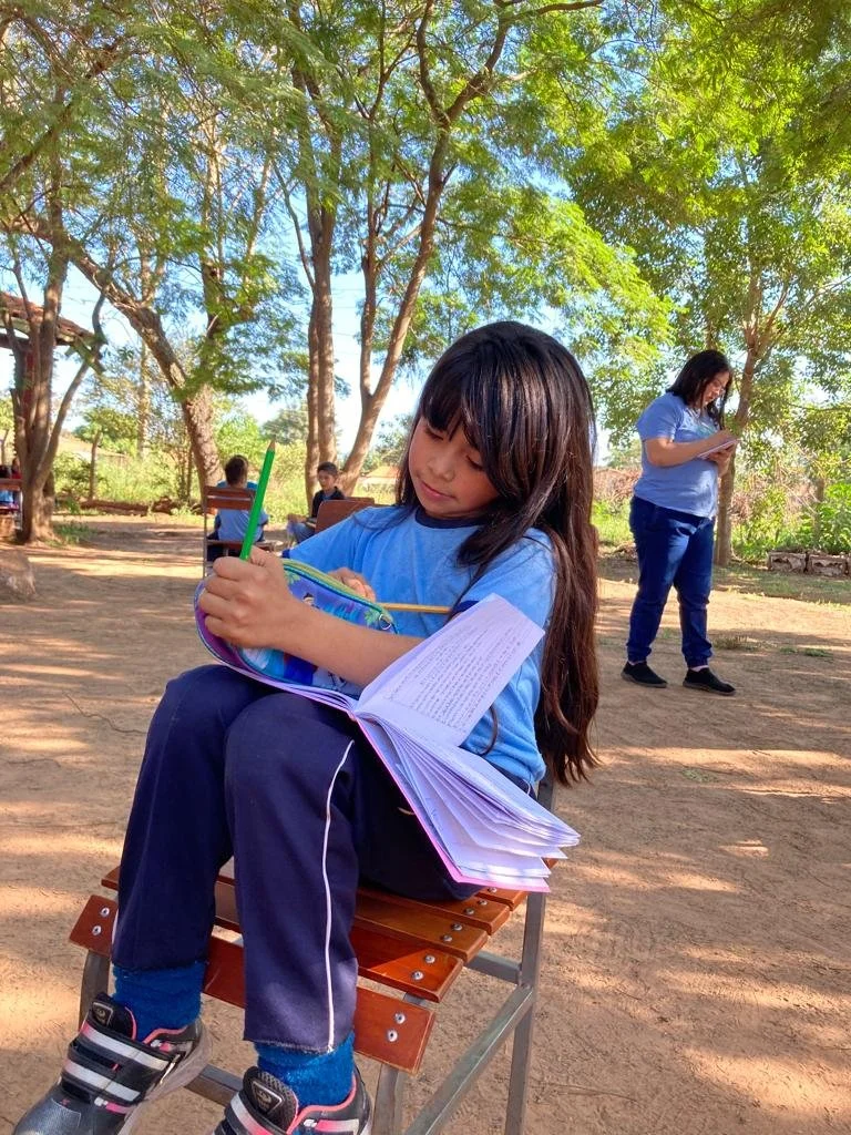 A girl sitting on a wooden bench outdoors, writing in a notebook, with a woman standing in the background reading a book, surrounded by trees and sunlight.