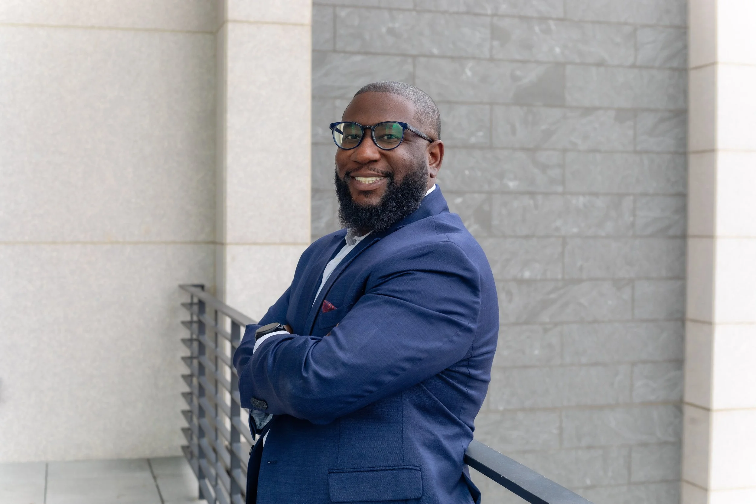 A man with glasses, a beard, and a smile, standing outdoors against a stone wall, wearing a blue suit with a white shirt, arms crossed.