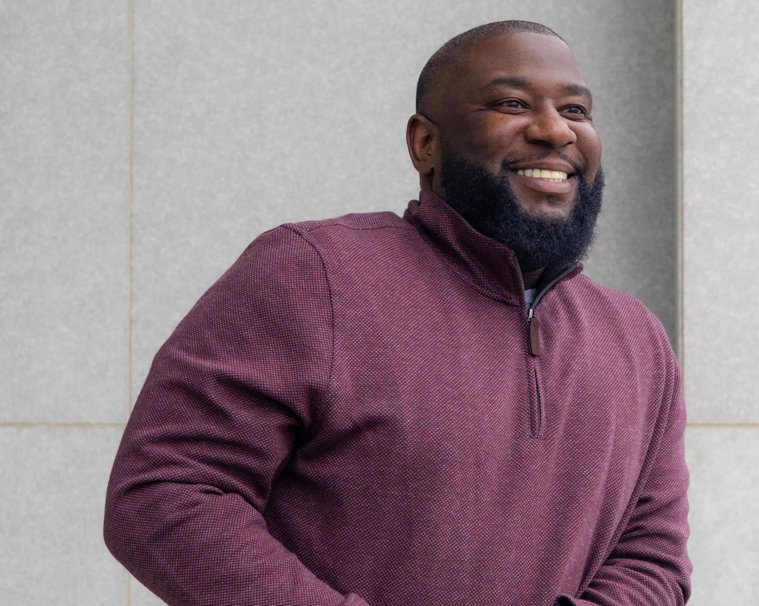 A smiling man with a beard wearing a maroon zip-up sweater, standing against a light gray tiled wall.