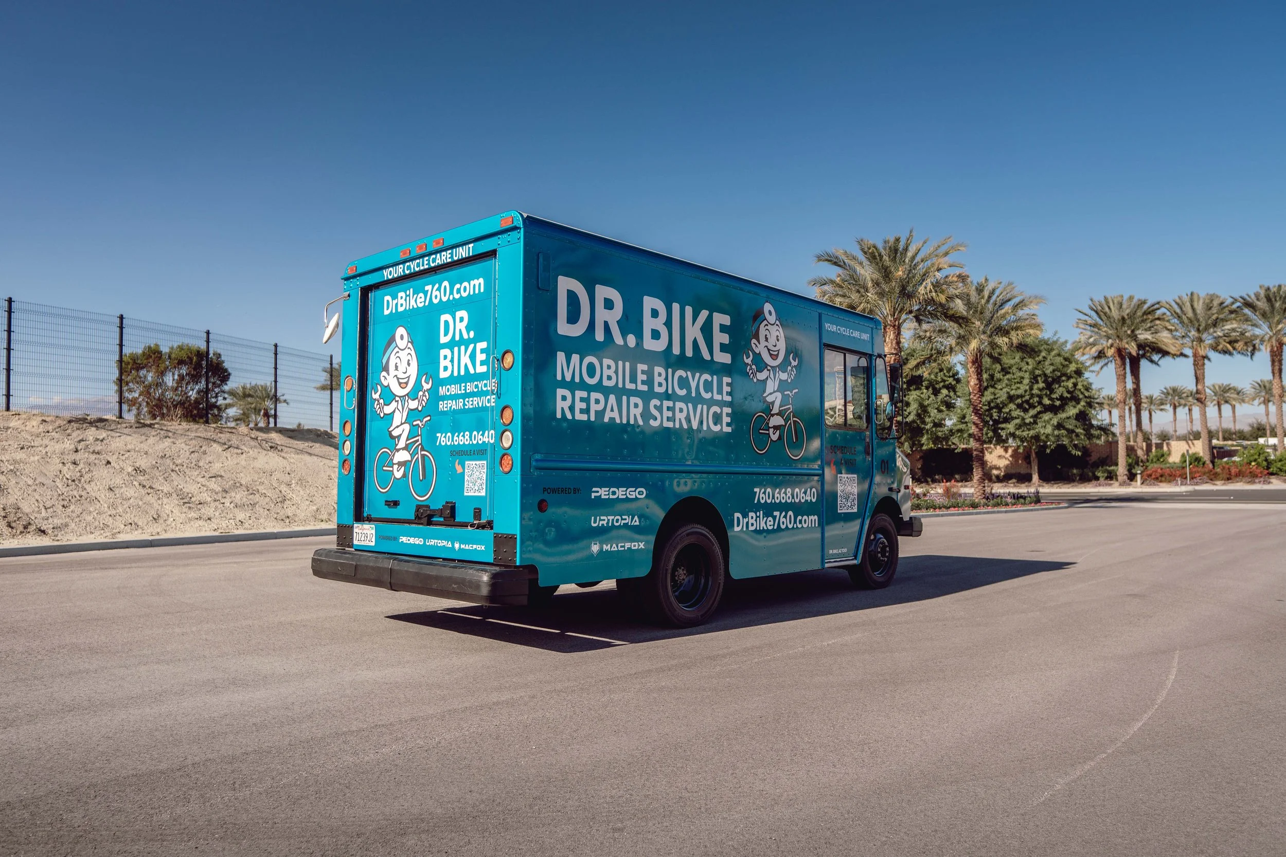 Blue mobile bicycle repair service truck on a paved road with palm trees and fenced area in the background.